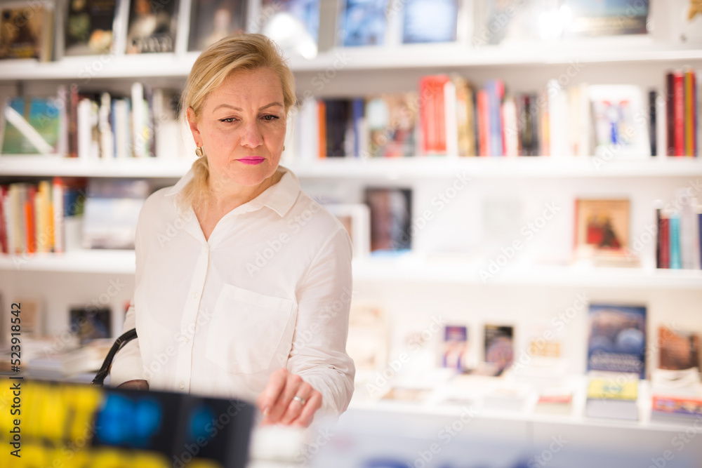 Portrait of handsome adult mature lady choosing new books on shelves in museum shop