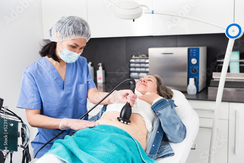 Confident female doctor performing radiofrequency lifting procedure on abdomen of mature woman