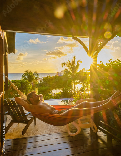 Fotografie, Obraz Young men in swim shorts sunbathing in a hammock at Saint Lucia Caribbean, men at infinity pool during sunset