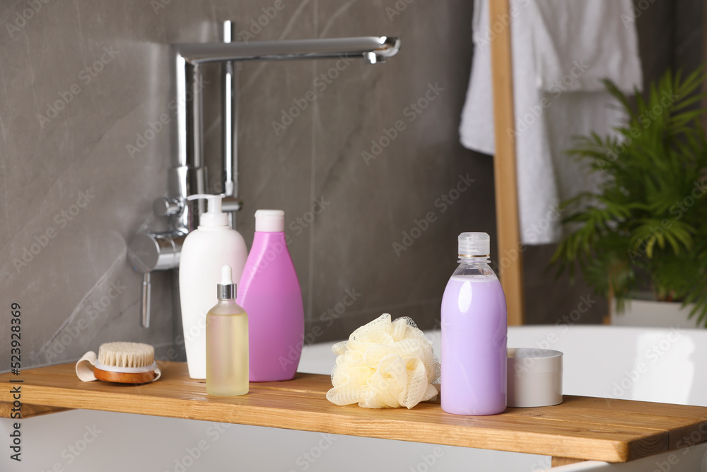 Wooden bath tray with shampoo, hair oil and other toiletries on tub indoors Wooden bath tray with shampoo, hair oil and other toiletries on tub indoors