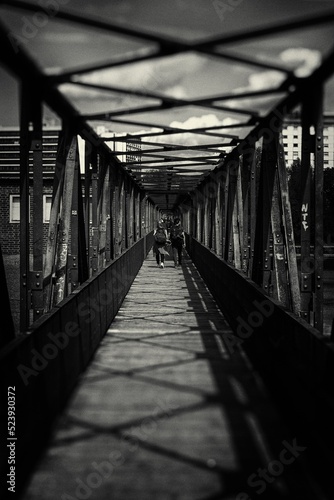 Vertical grayscale shot of a bridge with people walking in daylight with light and shadows on it photo