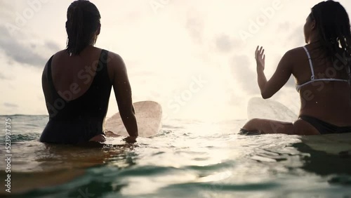  Low angle view of Asian woman surfer paddling surfboard and riding the wave in the sea at tropical beach at sunset. Girl friends enjoy outdoor lifestyle and water sport surfing on summer vacation