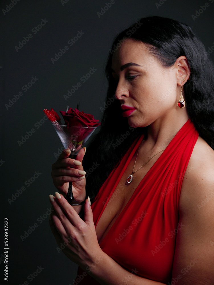 brunette with red lipstick on her lips in a red dress with red stones in jewelry with a red rose in a martini glass on a gray background