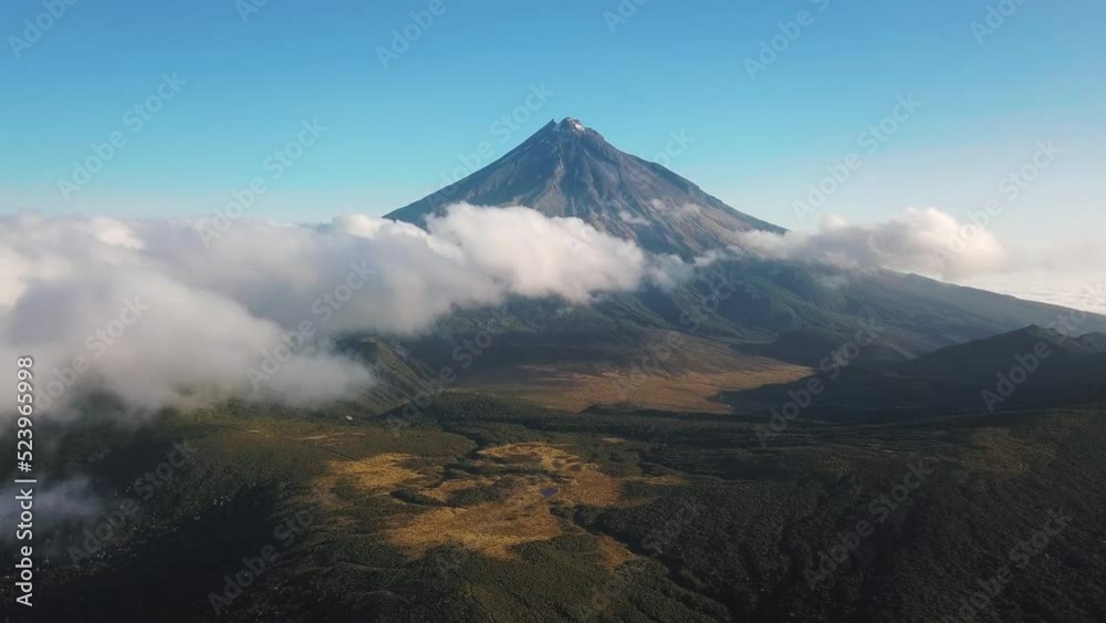 Mount Taranaki volcano behind white clouds. Passing over a forest valley. Mount Taranaki volcano behind white clouds. Passing over a forest valley.