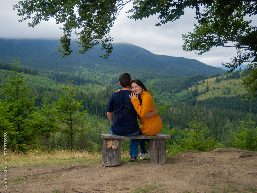Young man and woman admiring breathtaking view while sitting on bench in the mountains. Lovely couple in casual clothes sit under large tree with forest background. Young man and woman admiring breathtaking view while sitting on bench in the mountains. Lovely couple in casual clothes sit under large tree with forest background.