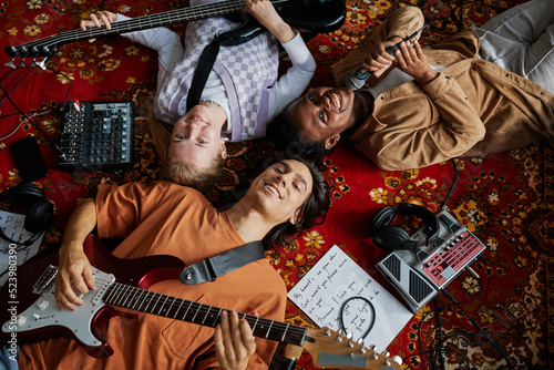 Top view shot of smiling music band lying on ornate carpet in mucis studio and looking at camera