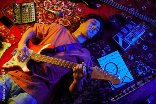 Top view shot of young musician playing guitar while lying on carpet lit by neon light photo