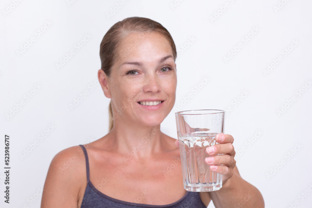 Caucasian beautiful smiling middle aged woman holding glass of pure water on white background looking at camera. Focus on glass Caucasian beautiful smiling middle aged woman holding glass of pure water on white background looking at camera. Focus on glass