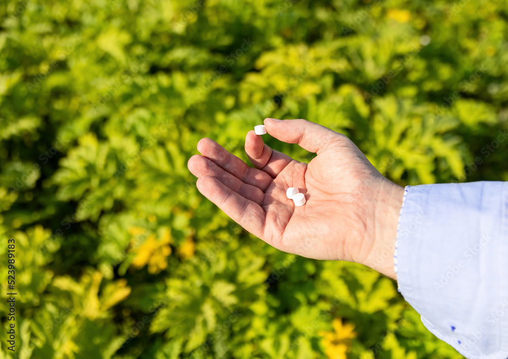 Hand holding white chemical pills against brightly lit green leaves background. 