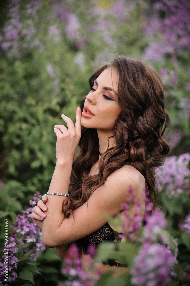 a beautiful girl with makeup and hair styling in underwear stands in a garden with lilacs on a summer evening a beautiful girl with makeup and hair styling in underwear stands in a garden with lilacs on a summer evening