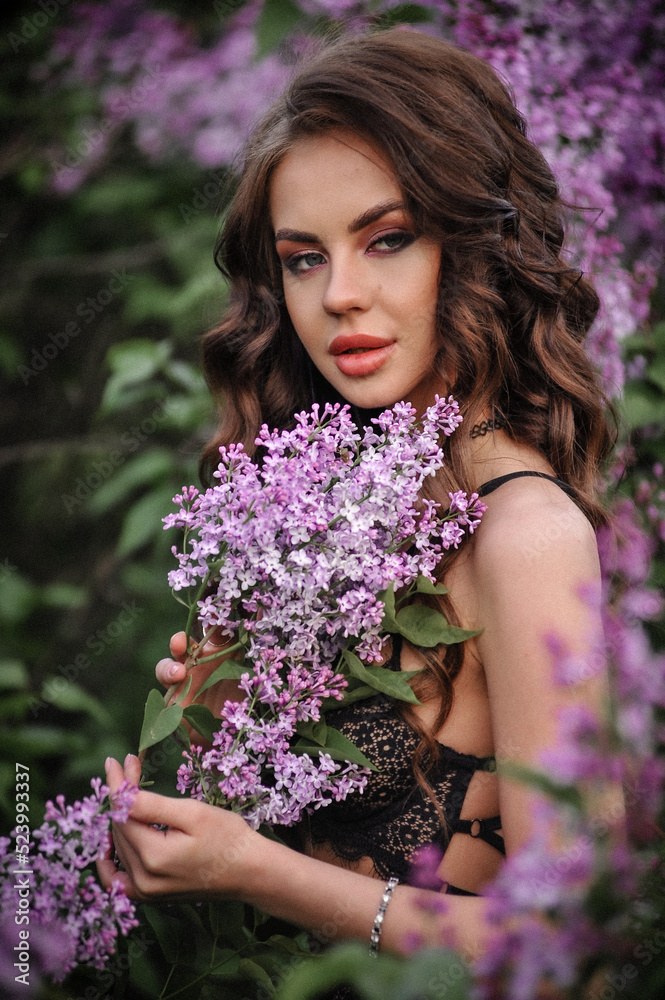 a beautiful girl with makeup and hair styling in underwear stands in a garden with lilacs on a summer evening a beautiful girl with makeup and hair styling in underwear stands in a garden with lilacs on a summer evening