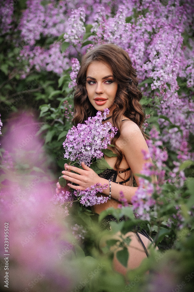 a beautiful girl with makeup and hair styling in underwear stands in a garden with lilacs on a summer evening a beautiful girl with makeup and hair styling in underwear stands in a garden with lilacs on a summer evening