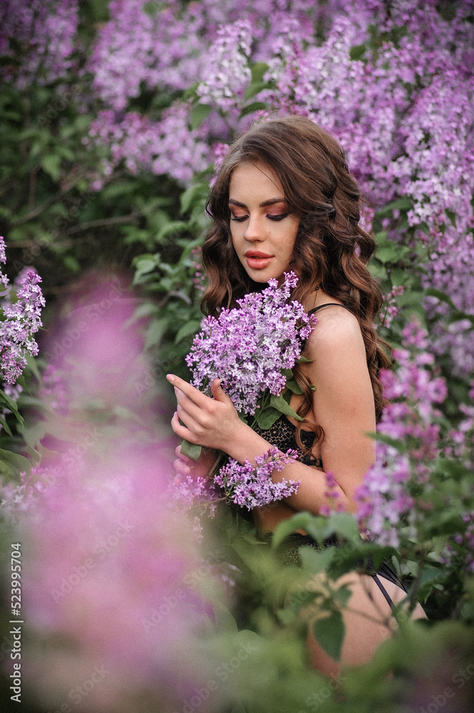 a beautiful girl with makeup and hair styling in underwear stands in a garden with lilacs on a summer evening