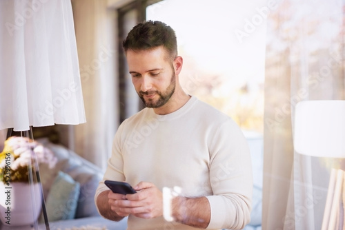 Handsoma man text messaging while standing at the window at home