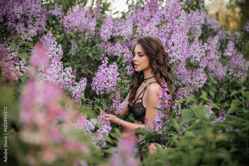 a beautiful girl with makeup and hair styling in underwear stands in a garden with lilacs on a summer evening a beautiful girl with makeup and hair styling in underwear stands in a garden with lilacs on a summer evening