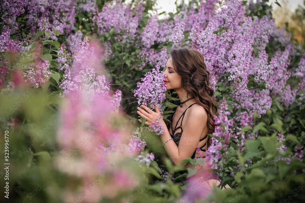 a beautiful girl with makeup and hair styling in underwear stands in a garden with lilacs on a summer evening
