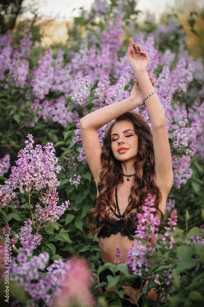a beautiful girl with makeup and hair styling in underwear stands in a garden with lilacs on a summer evening a beautiful girl with makeup and hair styling in underwear stands in a garden with lilacs on a summer evening
