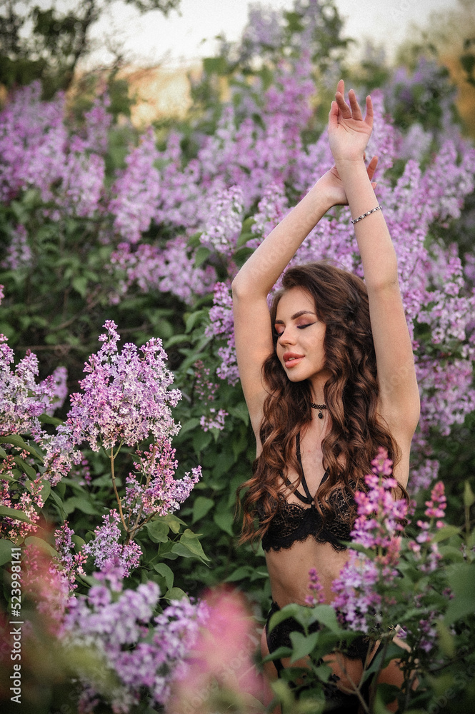 a beautiful girl with makeup and hair styling in underwear stands in a garden with lilacs on a summer evening