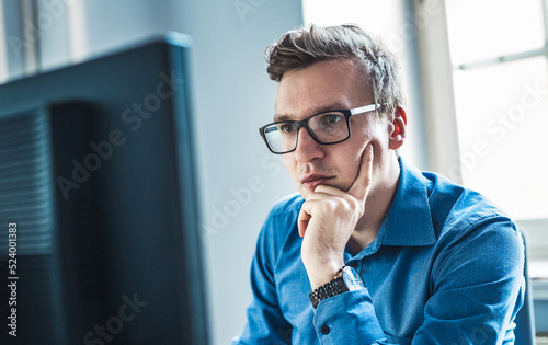Canvas Print Handsome Young Businessman Wearing Eyeglasses Sitting at his Table Inside the Office, Looking at the Report on his Computer Screen