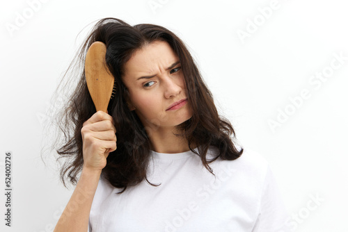 a sad, upset woman tries to comb her long, dark, tangled hair with a wooden massage comb, standing in a white T-shirt on a white background