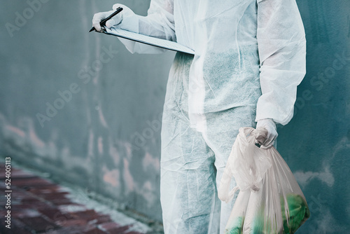 Forensic investigator collecting evidence on a murder scene on a street holding a plastic bag and wearing a hazmat suit. Crime researcher doing a scientific criminal investigation outdoors in a city photo