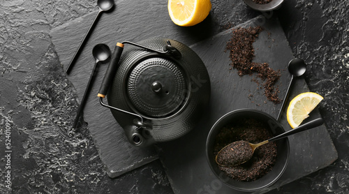 Teapot, bowl with dry tea leaves and lemon on dark background, top view
