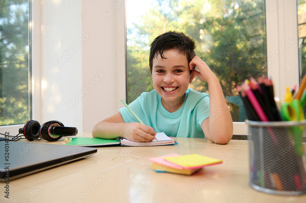 Portrait of a teenager school boy, student of an online school, smiling a sincere toothy smile while doing homework. Stationery , laptop with audio headset and school supplies lying on the desk Portrait of a teenager school boy, student of an online school, smiling a sincere toothy smile while doing homework. Stationery , laptop with audio headset and school supplies lying on the desk