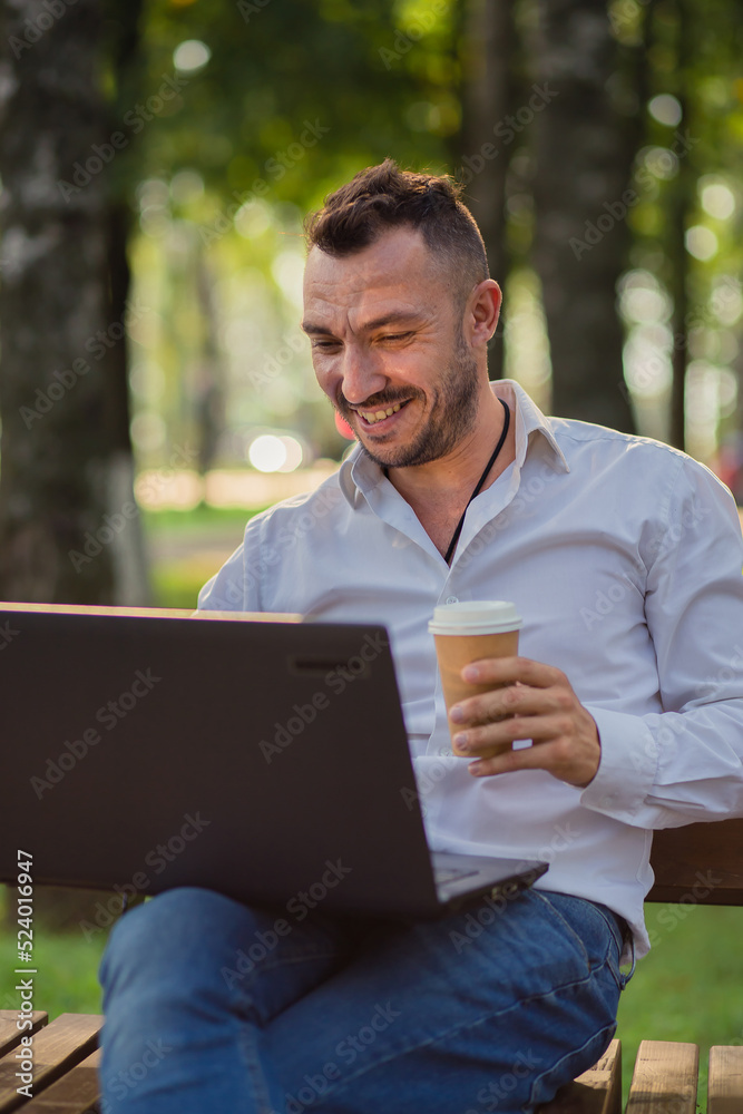 Smiling businessman is working in the park with a laptop, drinking coffee. A young man on a background of green trees, a hot sunny summer day. Warm soft light, close-up. Smiling businessman is working in the park with a laptop, drinking coffee. A young man on a background of green trees, a hot sunny summer day. Warm soft light, close-up.