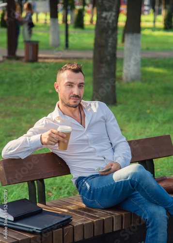 A pensive man sits on a bench, drinks coffee, holds dollars. A young man on a background of green trees, a hot sunny summer day. Warm soft light, close-up.