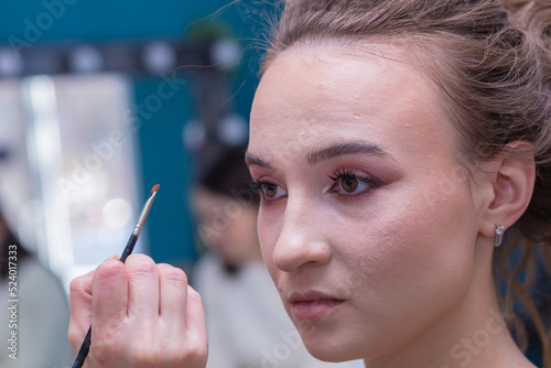 Makeup artist's hand with a brush near the girl's face in the salon. Female master makes makeup to a young woman. Business concept - beauty salon, facial skin care, cosmetology.
