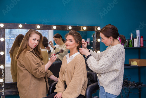 Two masters professionally style hair and make up a girl in the salon. Make-up artist and hairdresser create an image of a young woman. Business concept - beauty salon, facial skin and hair care.