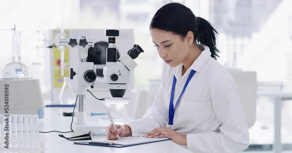 Young female scientist with microscope doing paperwork in science innovation for a clinical trial. Expert female using technical equipment to analyze DNA. Professional woman doing medical research.