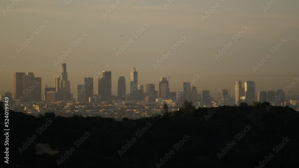 Downtown Los Angeles, California as seen from the mountains northwest of the city on a summer morning. An airplane flies over the iconic cityscape. A hazy grey sky hangs over the large metropolis.