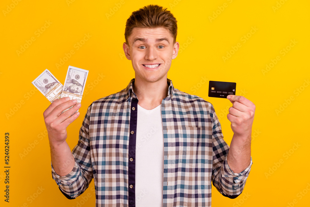 Photo of young cheerful man hold hand credit card cash savings investment isolated over yellow color background Photo of young cheerful man hold hand credit card cash savings investment isolated over yellow color background
