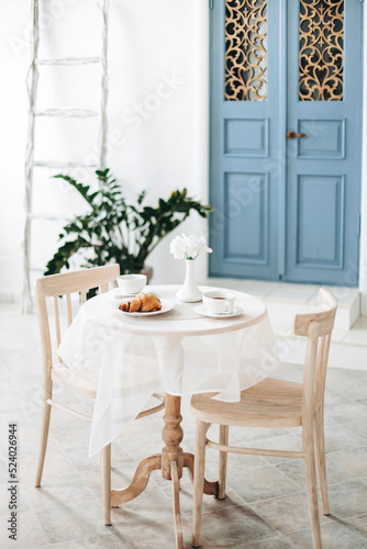 Romantic seating area with beige chairs and a round vintage table  against a blurred background of a white mediterranean house with a blue door and a potted plant near the wall. Patio of Santorini