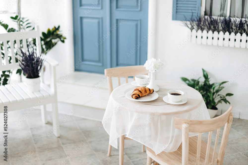 Romantic seating area with beige chairs and a vintage table with two cups of coffee and a croissant on a blurred background of blue door of a mediterranean white house Romantic seating area with beige chairs and a vintage table with two cups of coffee and a croissant on a blurred background of blue door of a mediterranean white house