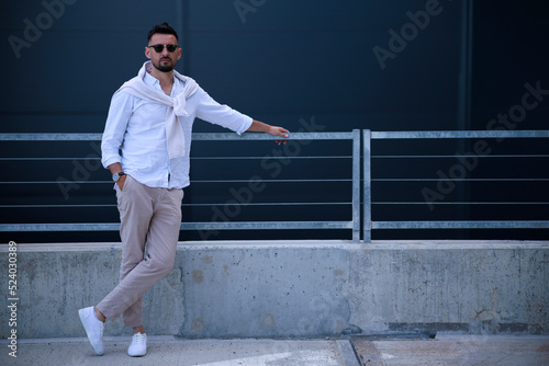 Cool modern young hipster man with a stylish hairstyle in sunglasses in a white shirt. Attractive urban guy posing near a dark wall. Fashionable spring menswear. © Евгений Бордовский