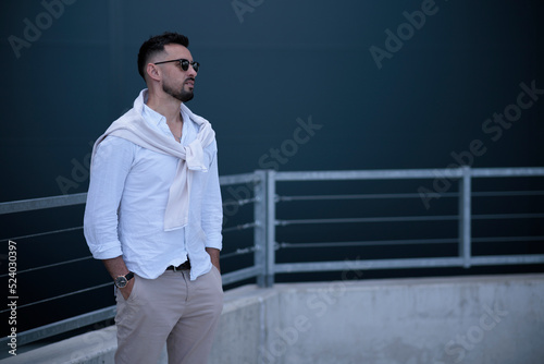 Young stylish brutal men in stylish clothes in sunglasses stands is in a city near a gray building. American guy. Fashionable men's summer clothes. Youth style.