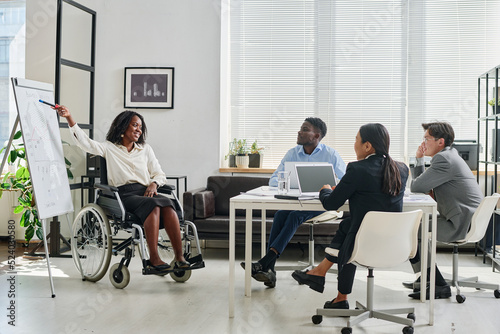 African businesswoman in wheelchair pointing at flipchart and presenting her report to colleagues during teamwork at office