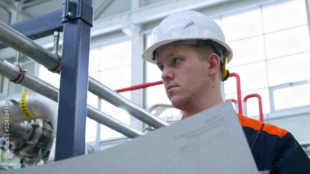 A young engineer reads the technological scheme of the equipment at the plant. Work in heavy industry with mechanical equipment. Worker in overalls and a white work helmet at the workplace. A young engineer reads the technological scheme of the equipment at the plant. Work in heavy industry with mechanical equipment. Worker in overalls and a white work helmet at the workplace.