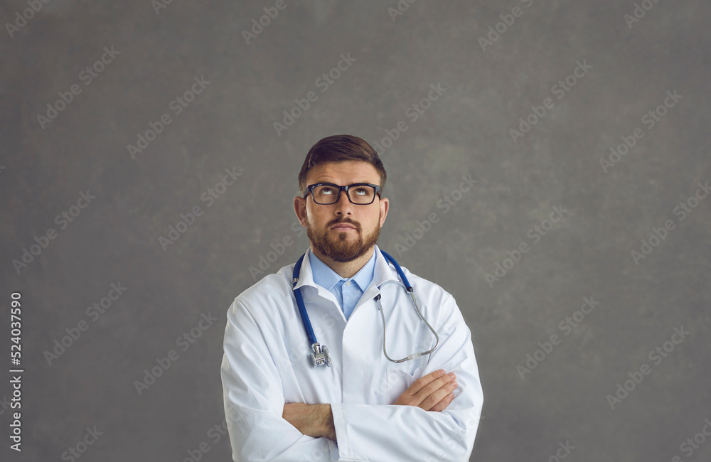 Studio portrait of a serious male doctor thinking about work. Young man in a white medical lab coat standing arms folded and looking up with a thoughtful face expression Studio portrait of a serious male doctor thinking about work. Young man in a white medical lab coat standing arms folded and looking up with a thoughtful face expression