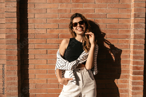 Smiling young caucasian woman posing looking at camera standing near wall in sunny weather. Brown-haired girl wears sunglasses, top, sweatshirt and pants. Happy weekend concept.