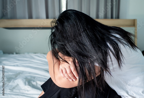 Asian woman in a white shirt sits with her head down on the bed, showing stress, disappointment, depression, migraines, health issues, health care and stress management concept.