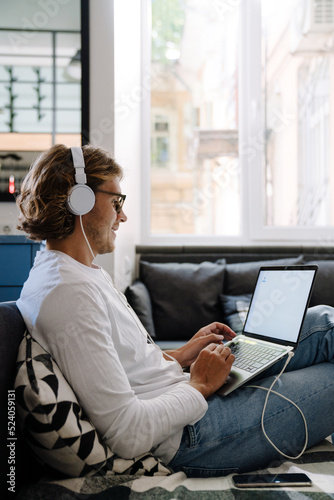 Young smiling long-haired man in glasses and headphones working laptop