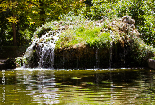 Artificial stone fountain on a small pond. Water flows in a lake  mountain river. A natural landscape in a green public state park  forest  square against green trees. Calm aqua in a water reservoir.