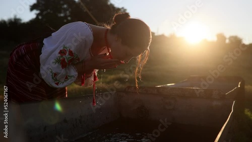 Young Ukrainian woman washes her face in traditional national embroidered shirt and skirt on meadow at sunset.
