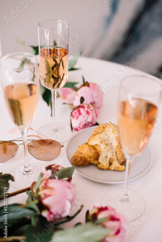 Delicious rose wine in glasses, pink peonies and a croissant on the white table background, flat lay, top view
