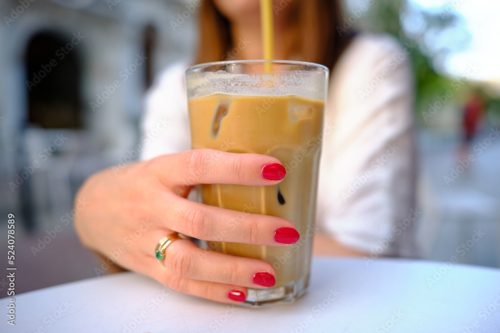Iced coffee with milk. Close-up of a woman's hand holding a coffee with a straw and ice. Hand with red nails. Iced coffee being drunk in a cafe outside on a summer sunny day. Iced coffee with milk. Close-up of a woman's hand holding a coffee with a straw and ice. Hand with red nails. Iced coffee being drunk in a cafe outside on a summer sunny day.