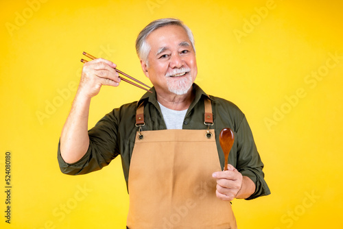Portrait charming retired asian elder man has a white mustache and beard wearing apron holding fork spoon yellow background.