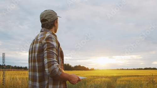 Farmer with a tablet computer in front of a sunset agricultural landscape. Man in a countryside field. Country life, food production, farming and technology.